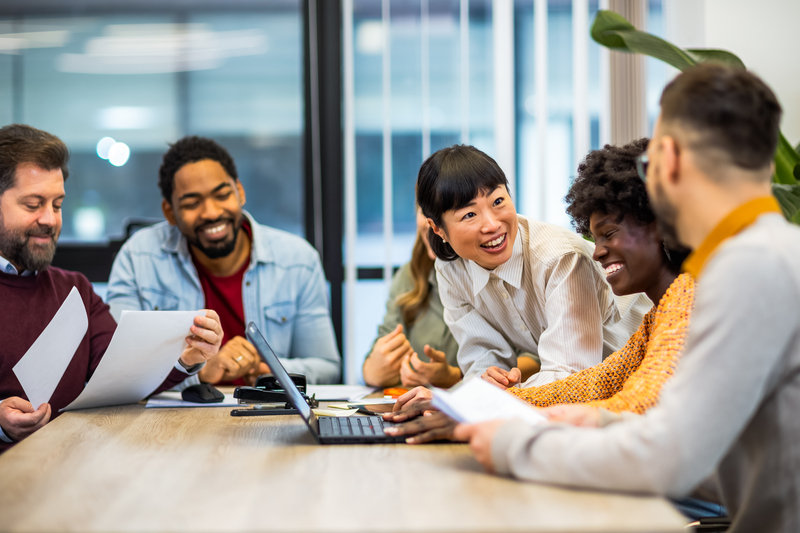 A diverse team gathers around a table, smiling and collaborating, representing the positive impact of using uplifting language in the workplace